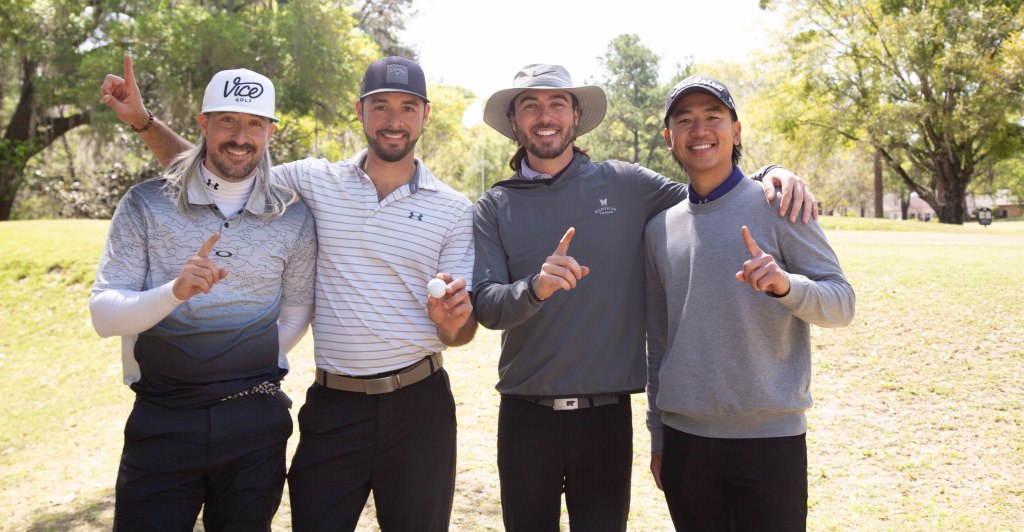Four men standing on a golf course, all smiling and holding up one finger, signifying a celebration or achievement.